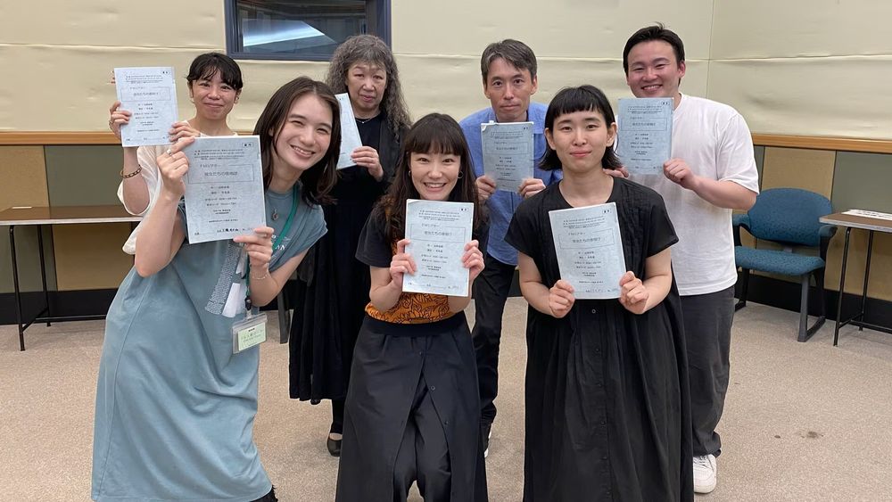 屋内で撮影された写真。７人の大人全員が台本を両手で持ってカメラ目線で笑顔。
