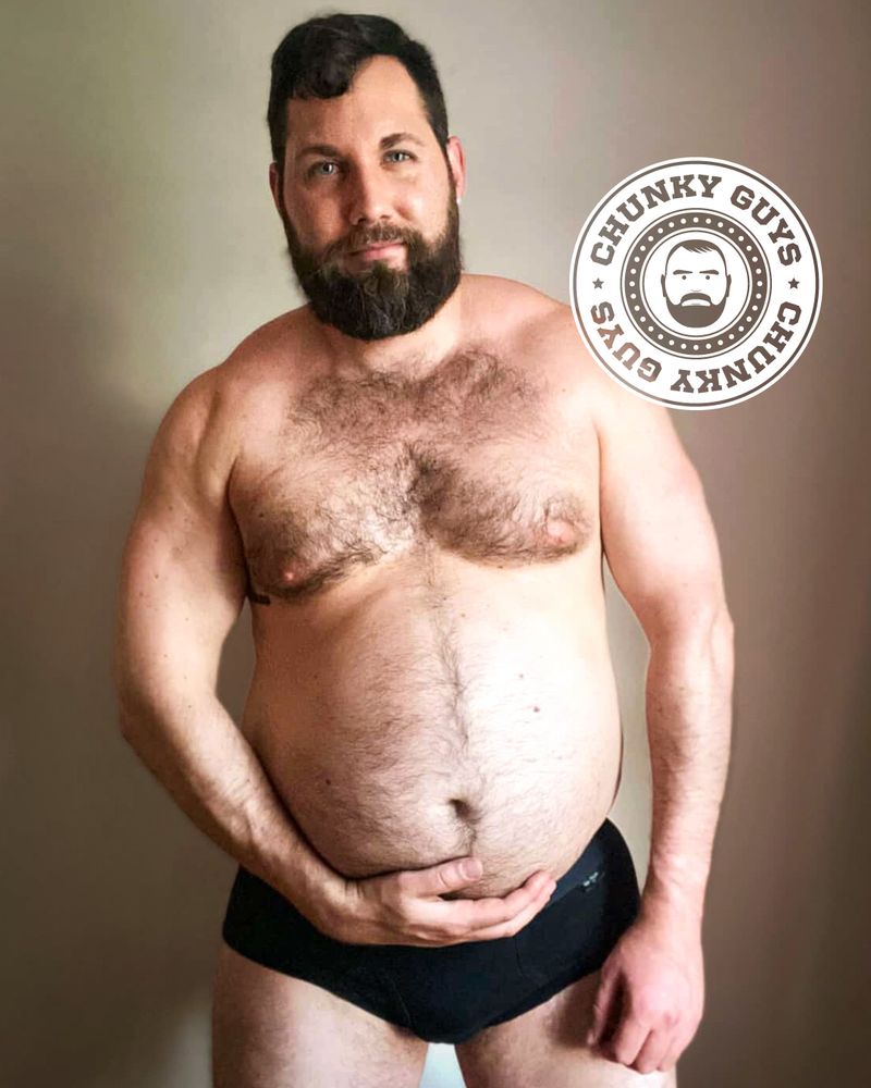 Bearded man with short dark hair standing against a plain background, wearing only black briefs. He has a hairy chest and a soft, round belly, which he is holding with one hand while his other arm rests at his side. He is looking at the camera with a slight smile. A circular logo reading “Chunky Guys” with a bearded face graphic is placed near his shoulder.