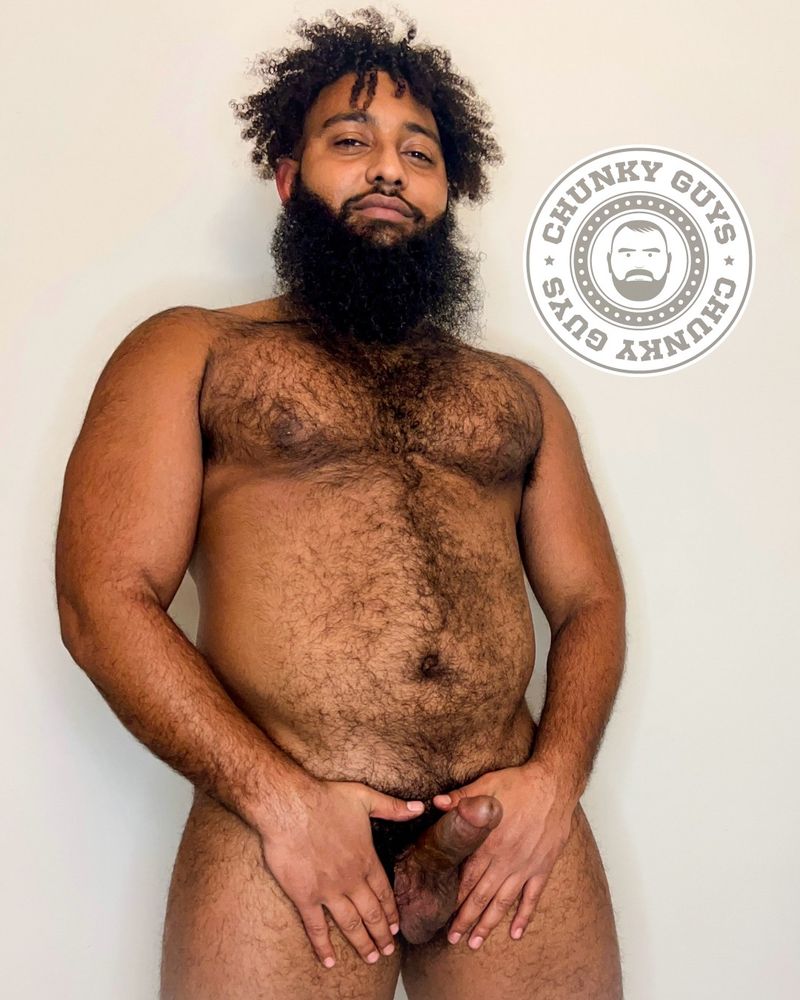 This photo features a bearded man with curly hair posing confidently against a plain background. He is naked and has a full, well-groomed beard and dark, curly hair that falls naturally. His expression is calm and self-assured, with a slight hint of a smile. He has a natural, hairy chest and an overall strong, solid physique. His hands are placed either side of his groin, showcasing his weighty balls and hard cut cock that is proudly pointing upward.

To the left of the man, there is a circular logo with the text "CHUNKY GUYS" repeated around the border, enclosing an illustration of a bearded face. The logo complements the image's theme, reinforcing the branding of the "Chunky Guys" identity.

The lighting in the image is soft and even, highlighting the subject's natural features. The overall composition is simple but effective, with the subject centered and the logo positioned strategically to add context.