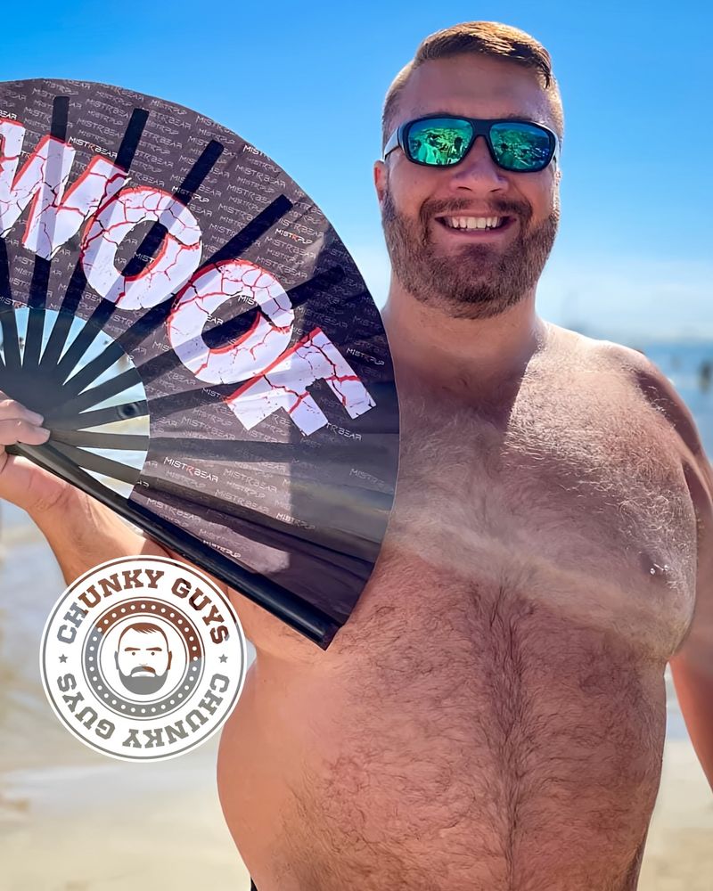 A smiling, bearded man with short brown hair stands shirtless on a sunny beach, wearing reflective sunglasses. He is holding an open black fan printed with the word “WOOF” in large, cracked white letters with red accents. A circular “CHUNKY GUYS” logo with a bearded face icon appears in the bottom left corner of the image. The blue sky and ocean are visible in the background.