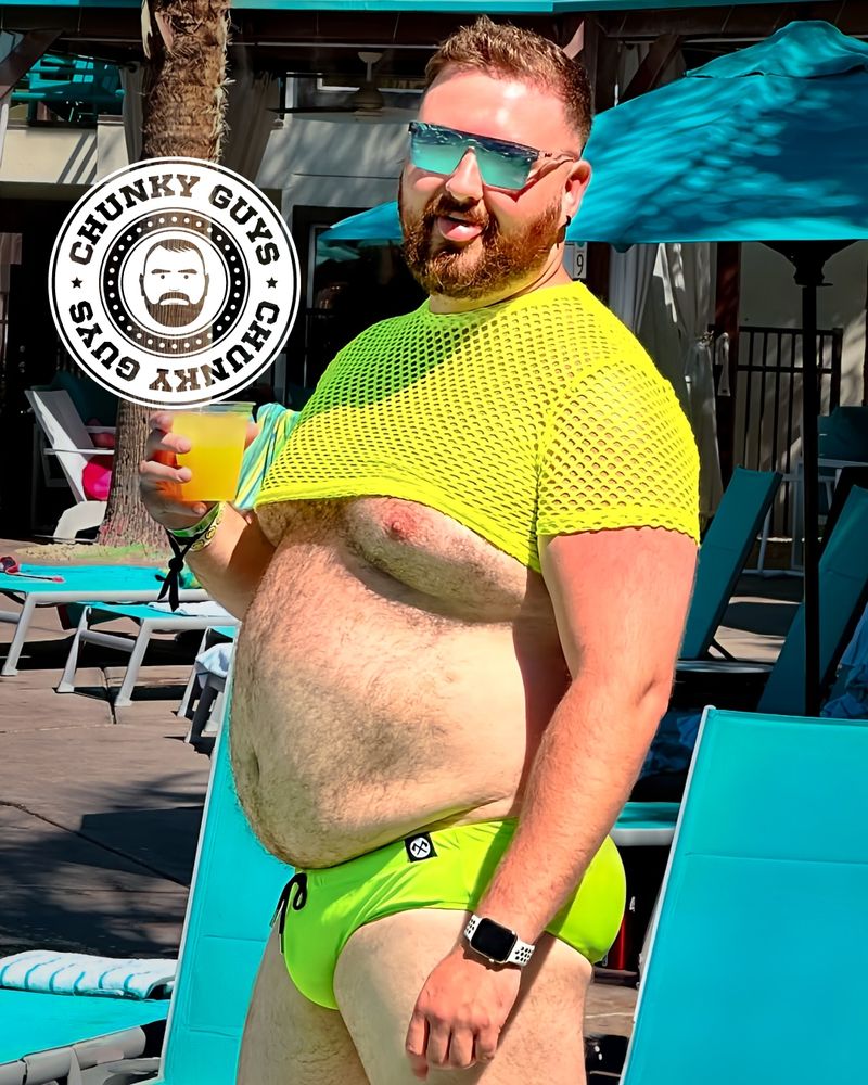 A bearded, plus-size man with sunglasses poses confidently by a pool, wearing a neon yellow mesh crop top and matching swim briefs. He holds a yellow drink in one hand and sticks out his tongue playfully. The "Chunky Guys" logo is overlaid near his head. The background features turquoise lounge chairs, umbrellas, and a palm tree.