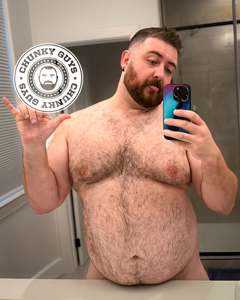 Bearded, plus-sized man taking a nude mirror selfie in a bathroom, holding his phone in one hand and making a playful gesture with the other. He has short dark hair, a trimmed beard, and a hairy chest and belly. A “Chunky Guys” logo has been added over part of the image.