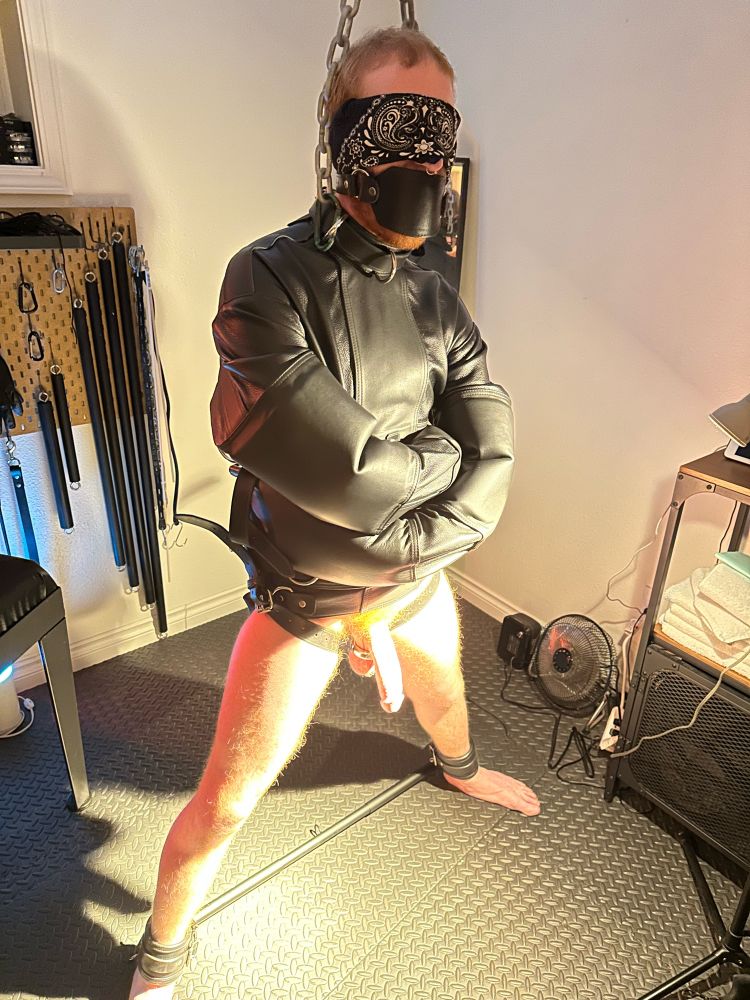 a naked person wearing a straitjacket, blindfolded, gagged, with a spreader bar keeping their legs apart.