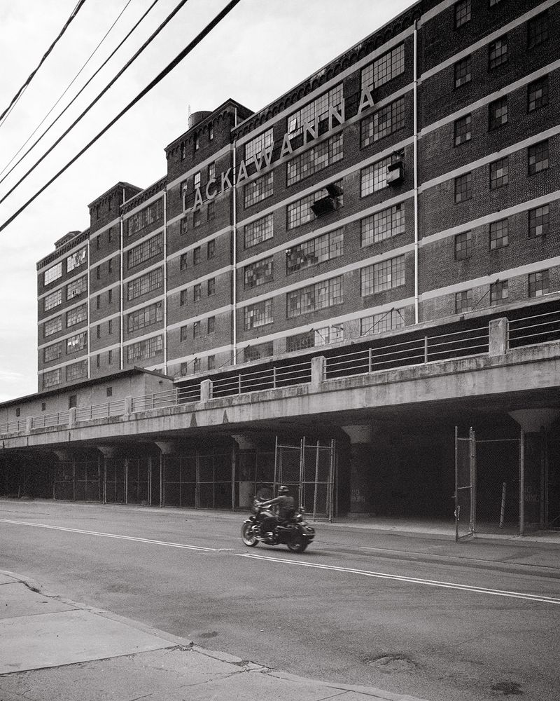 A man rides a motorcycle past a large, old warehouse building. The building says LACKAWANNA in metal letters suspended from the brick walls. 