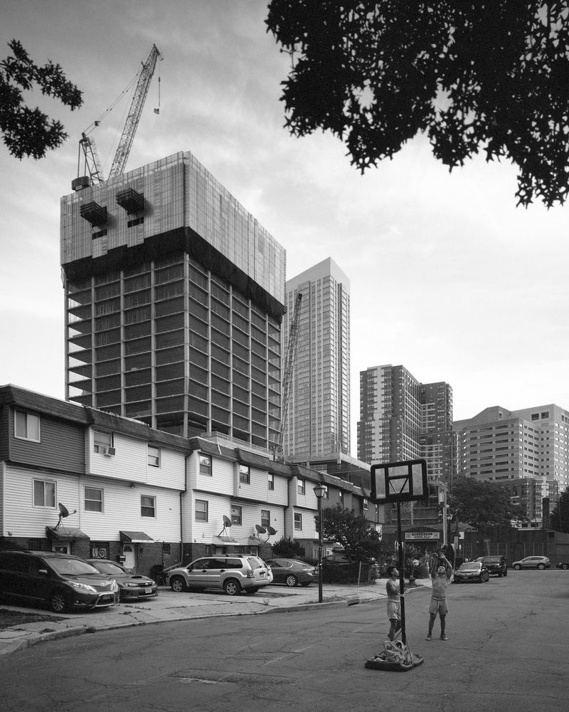Two teens shooting hoops on a cul-de-sac lined with low-rise two family homes. A residential tower is under construction in the background in front of other recently completed towers.