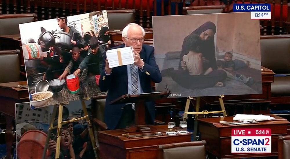 Bernie Sanders holds an approx. 6" stack of papers with the names of children who were murdered in Gaza on the senate floor with pictures of desperate hungry children behind him