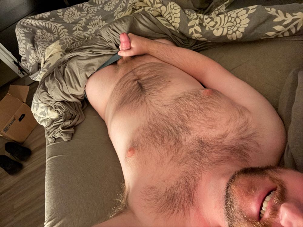 A naked hairy man laying in bed holding his erect cock while his smile peeks through
