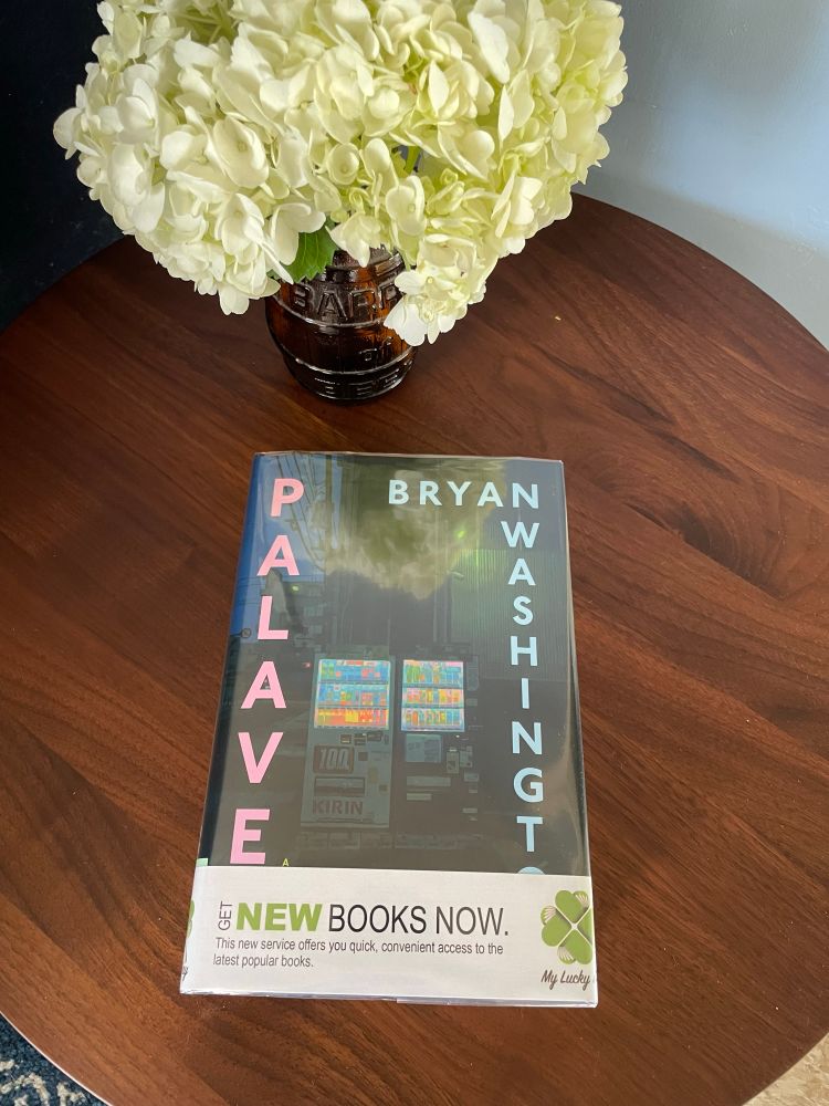 Photo of a book on a wooden table next to a vase with hydrangeas. The book is Palaver by Bryan Washington and shows a vending machine in Toyko at night. 