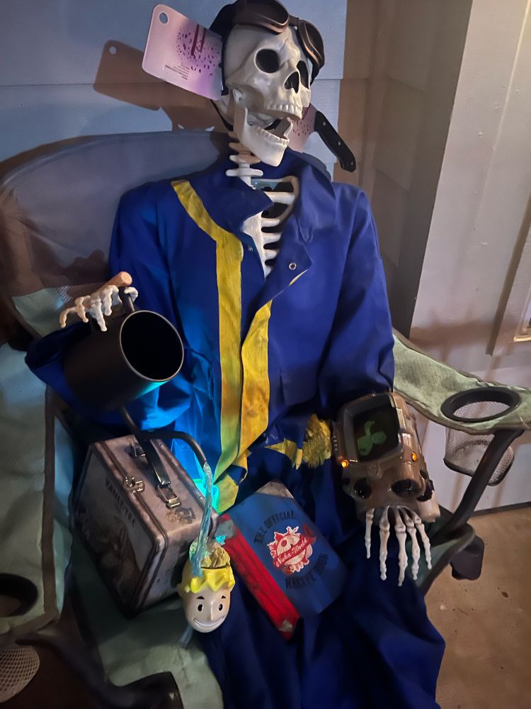 Photo of a Halloween prop skeleton sitting on a lawn chair wearing a Fallout Vault suit and Pip Boy, a costume hatchet through his head and goggles who is watering a vault boy planter with “brain fungus” and a vault Tec lunchbox next to him and a notebook on his lap that reads “the official Nuka World Recipe book”  with a faint glow coming from behind the lunch box and planter. 