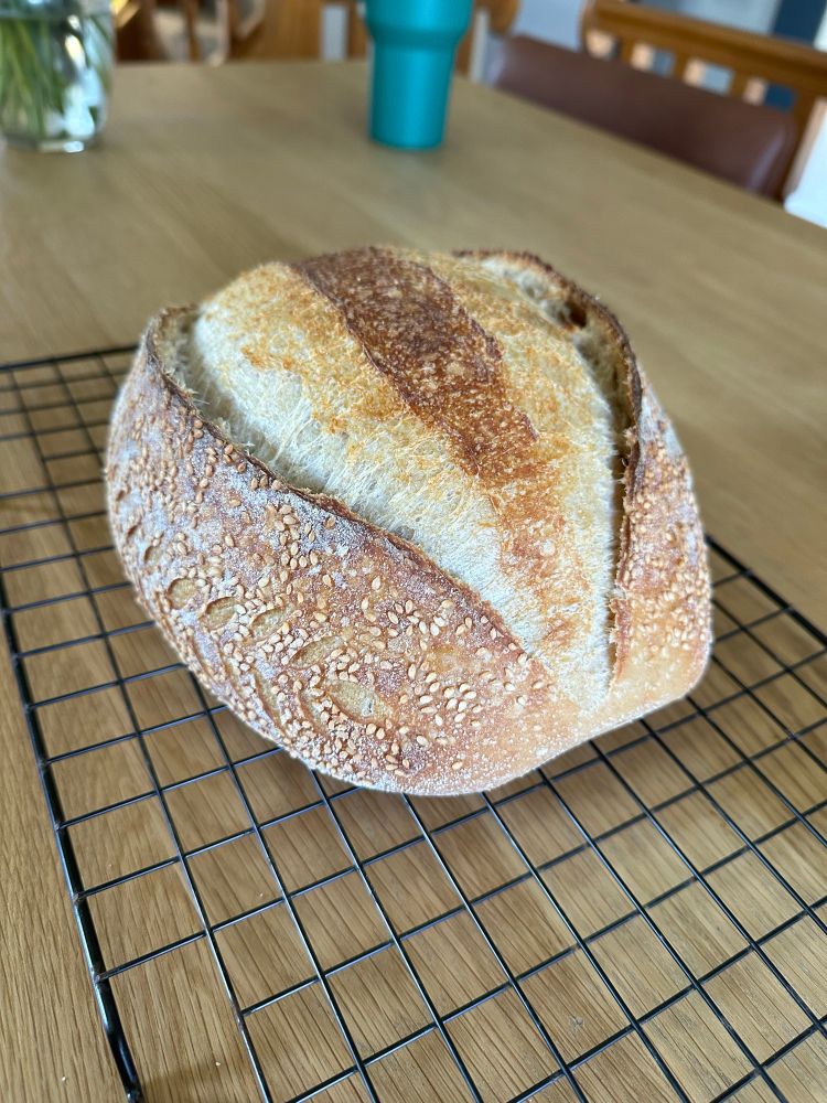Rustic sourdough loaf topped with sesame seeds, scored down the center, with leaf pattern on the sides 