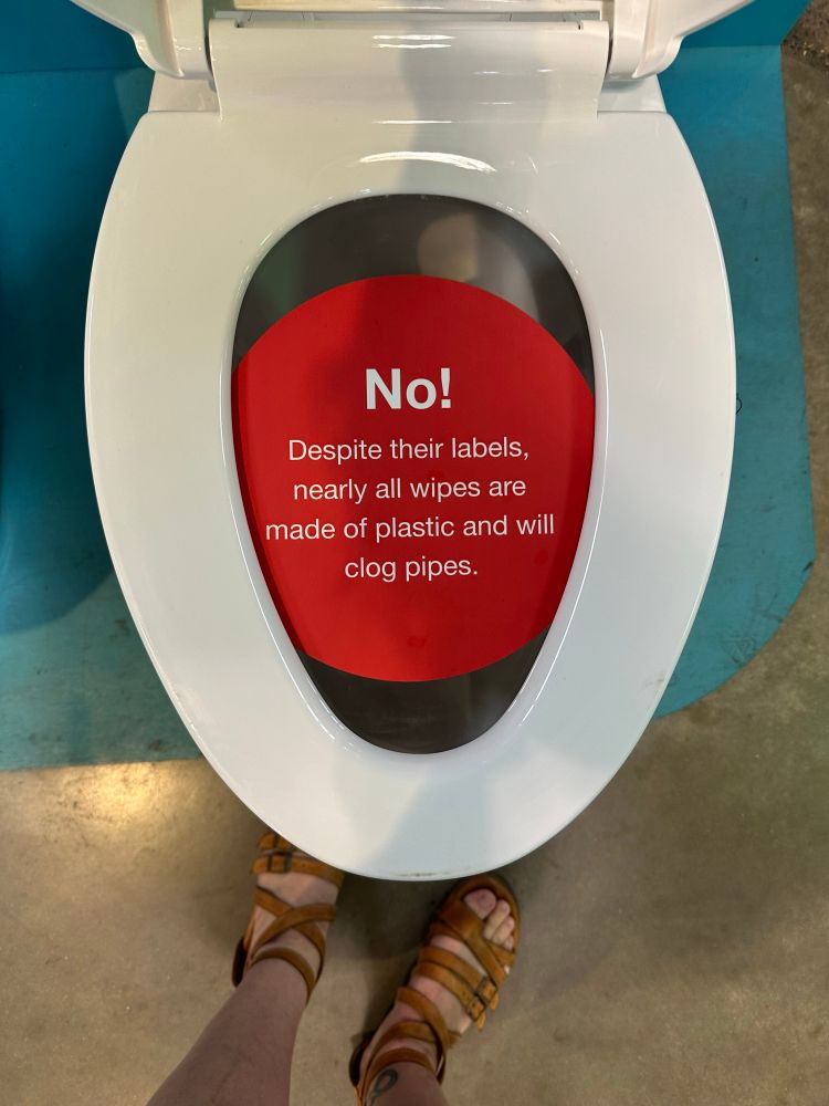 A toilet that is part of a display about flushable wipes from the Eco Building at the MN State Fair. The lid is up and a red label over the seat says, No! Despite their labels, nearly all wipes are made of plastic and will clog pipes.