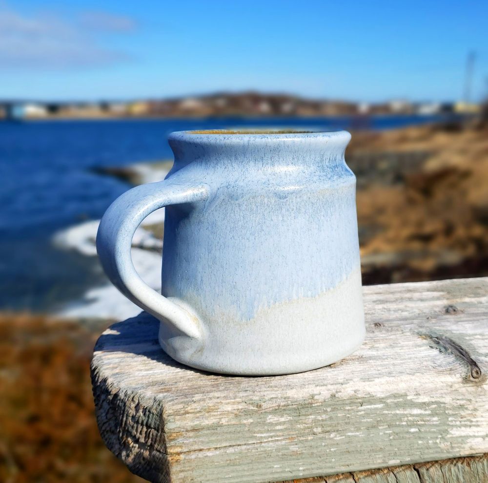 A pale blue mug sitting on a rough wooden board with a shoreline behind it and a blue sky overhead.