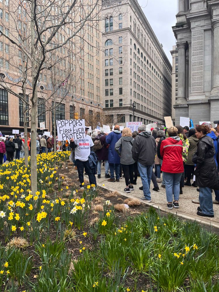 A large crowd of protestors mostly facing away from the camera as they round Philadelphia City Hall. There is an older white man standing near a flower bed of daffodils, holding a sign saying “Pop pop’s first ever protest”.