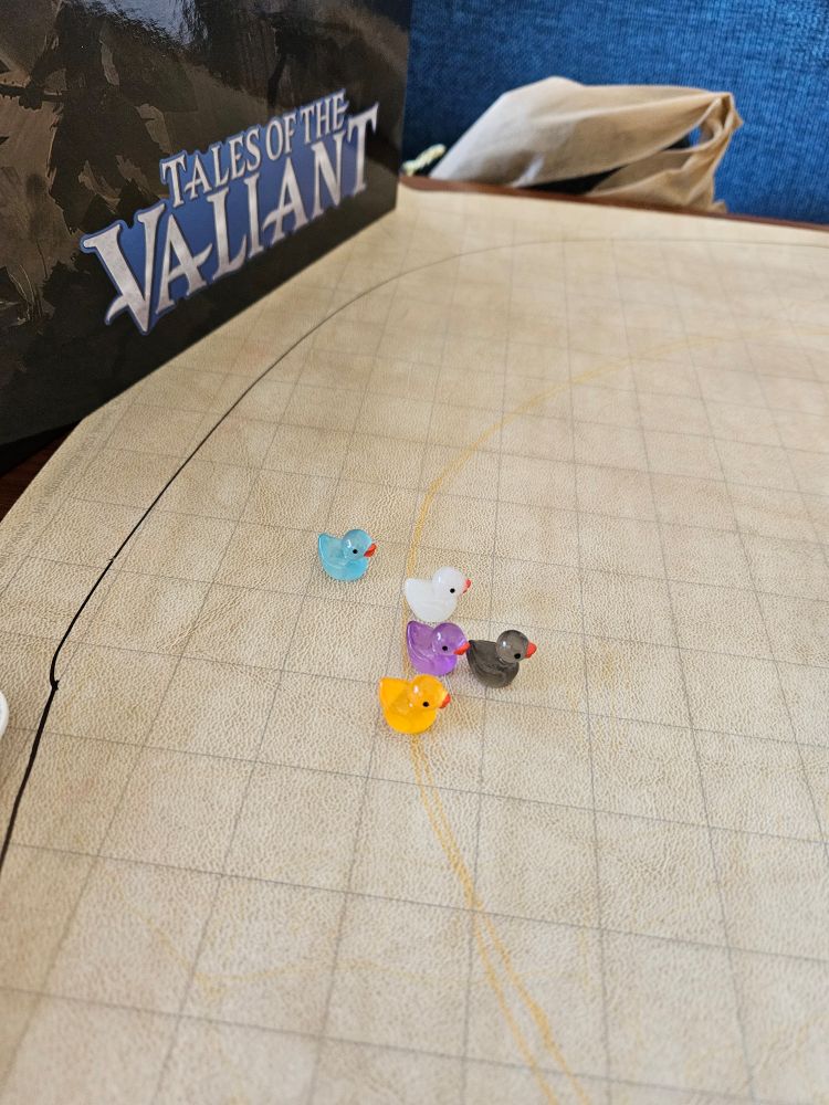 A table top rpg grid map has 5 variously coloured ducks as stand ins for character minis. A stand in the back showcases the game's name: Tales of the Valiant. 