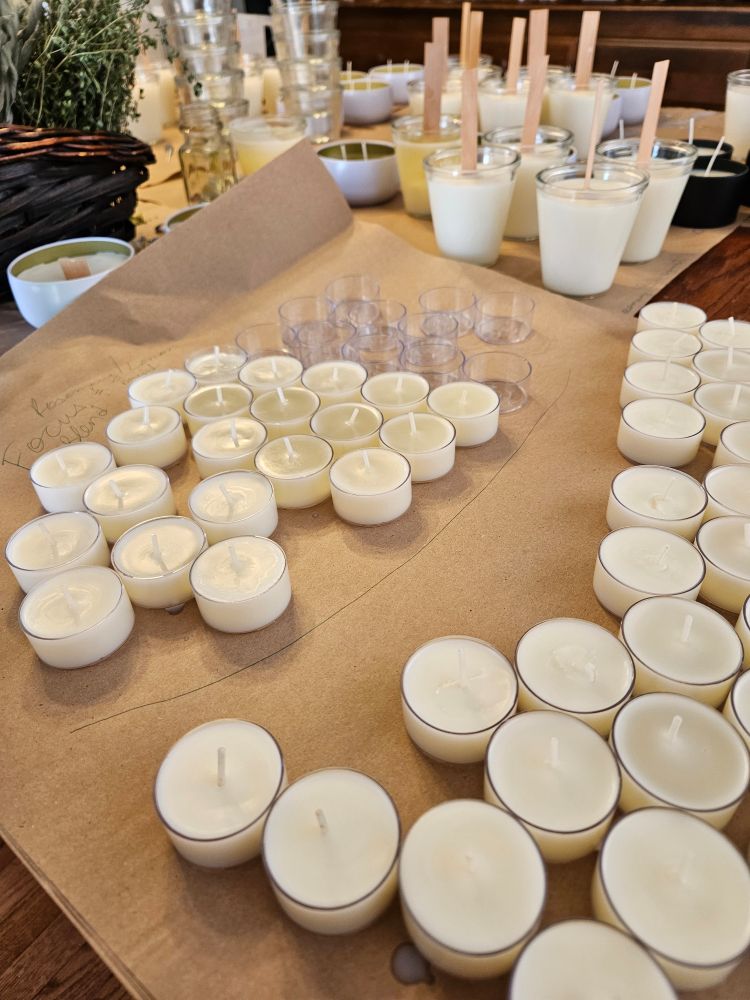 A tabletop candle-making workspace is covered in brown kraft paper, with rows of white tealight candles in both metal and clear plastic holders. To the left, several glass containers hold white wax with wooden wicks, mid-set. Behind them, more jars and vessels await use. A woven basket holds green herbs.