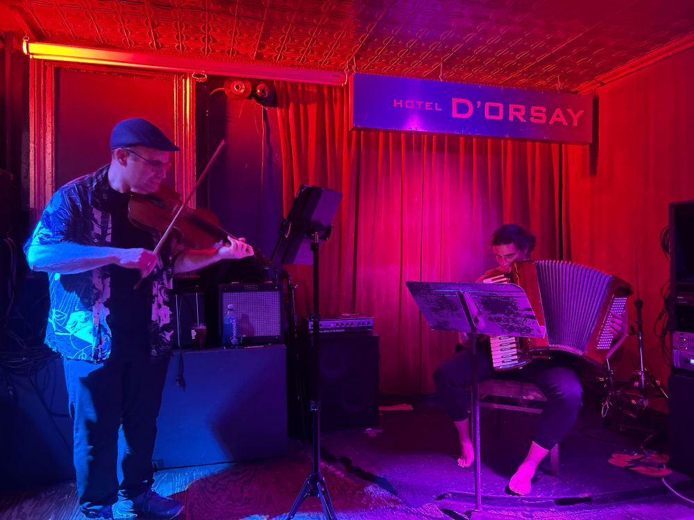 Ljova and Simone Baron performing at Barbes in Brooklyn. Ljova plays a fadolín (six string violin) and Simone plays an accordion. The background is dark red. 