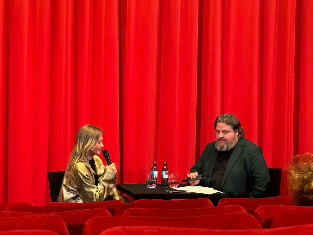 Das Bild zeigt zwei Personen, Julia Jentsch und einen Mitarbeiter des Filmmuseums, die auf einer Bühne sitzen, vor einem roten Vorhang. Die Person links, eine Frau mit langen blonden Haaren, hält ein Mikrofon in der Hand und scheint zu sprechen. Sie trägt eine goldene Jacke. Die Person rechts, ein Mann mit Bart und langen Haaren, sitzt ebenfalls mit einem Mikrofon in der Hand und scheint aufmerksam zuzuhören. Auf dem Tisch zwischen ihnen stehen zwei Flaschen und ein Glas. Im Vordergrund ein paar Sitzreihen des Kinosaals.
Aufgenommen beim fast einstündigen Gespräch vor dem Film.

Bereitgestellt von @altbot, privat und lokal generiert mit Ovis2-8B, etwas überarbeitet.