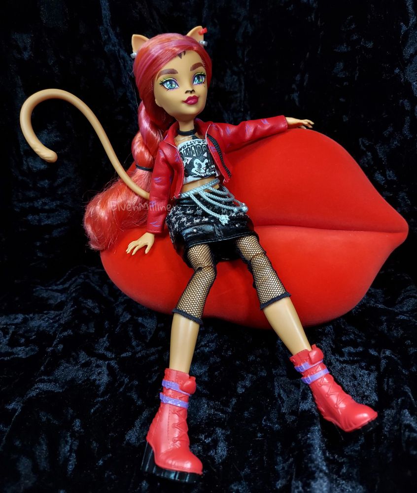 Monster High G3 Toralei Stripe core doll. Seated on Bratz red lips couch. Hair is in a big twist to her right shoulder (viewer's left). Wearing 3 small hoop earrings (red and silver), black choker necklace with a claw mark pendant, red pleather jacket with purple claw marks, white tube top with an illustration of a cartoon cat and the word "Hollywood", silver belt draped over black and silver houndstooth skirt, black fishnets ending at the calf, and red combat boots with purple stripes and black soles. Black crushed velvet background.