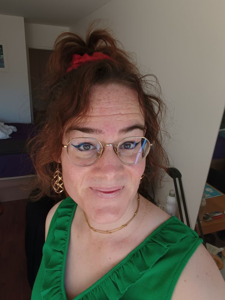 Selfie dans la même tenue, on voit un fin collier doré, des boucles d'oreille dorées aussi, faites de torsades, eyeliner assez voyant, cheveux rassemblés au-dessus de la tête avec un gros chouchou rouge, et deux mèches gardées pour encadrer mon visage