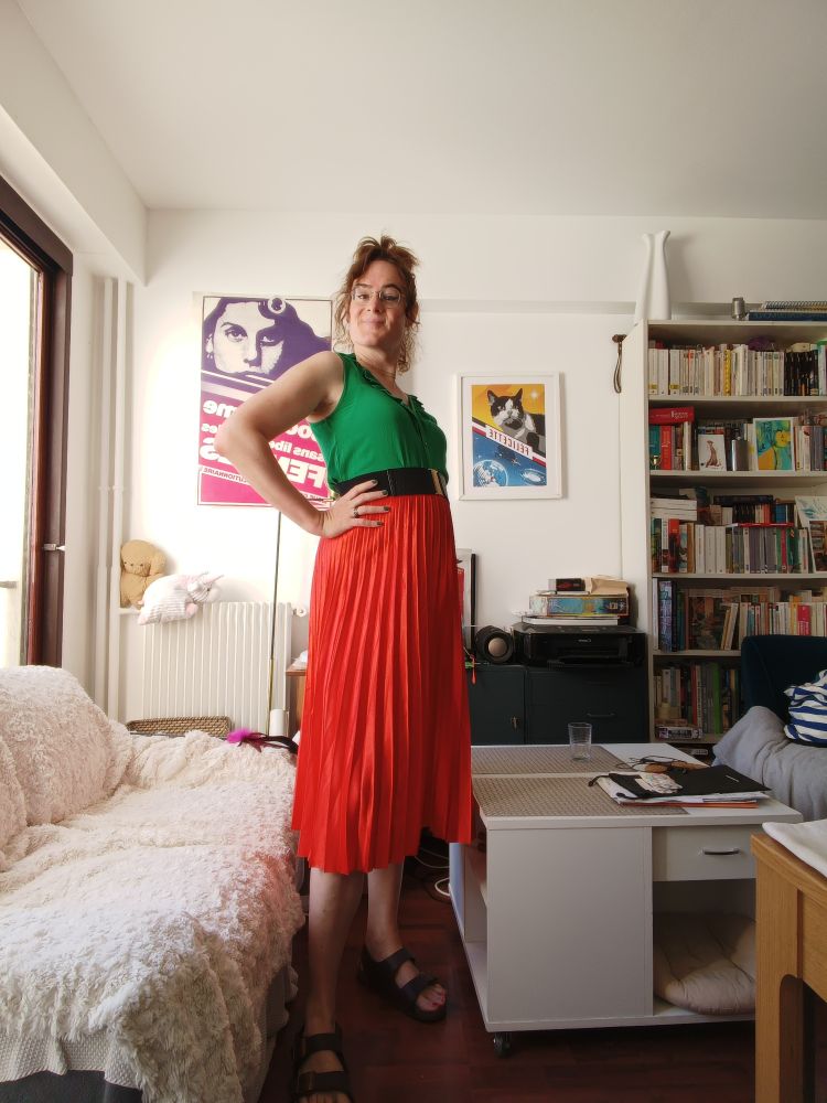 Photo de moi en pied dans mon salon, je porte des Birkenstock ocre, une jupe longue plissée orange, un haut vert sans manches, on voit des poils sous les bras parce que nique la peau lisse, j'ai la main gauche posée sur la hanche, on y voit une bague noire et du vernis vert sapin