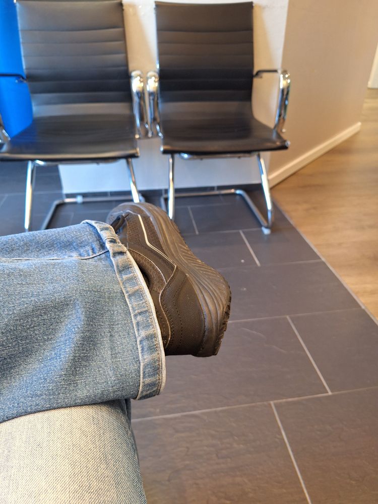 Fußboden einer Praxis mit zwei leeren  schwarzen Stühlen, davor mein übergeschlagenes Bein in Jeans und mit nem schwarzen Turnschuh. 