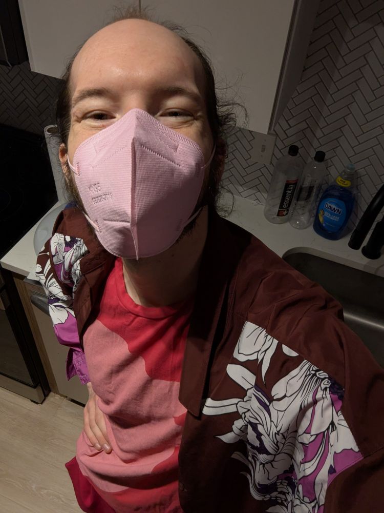 cheesing at the camera with a pink mask on on my kitchen. pink twitter bird shirt and flowery light long sleeves