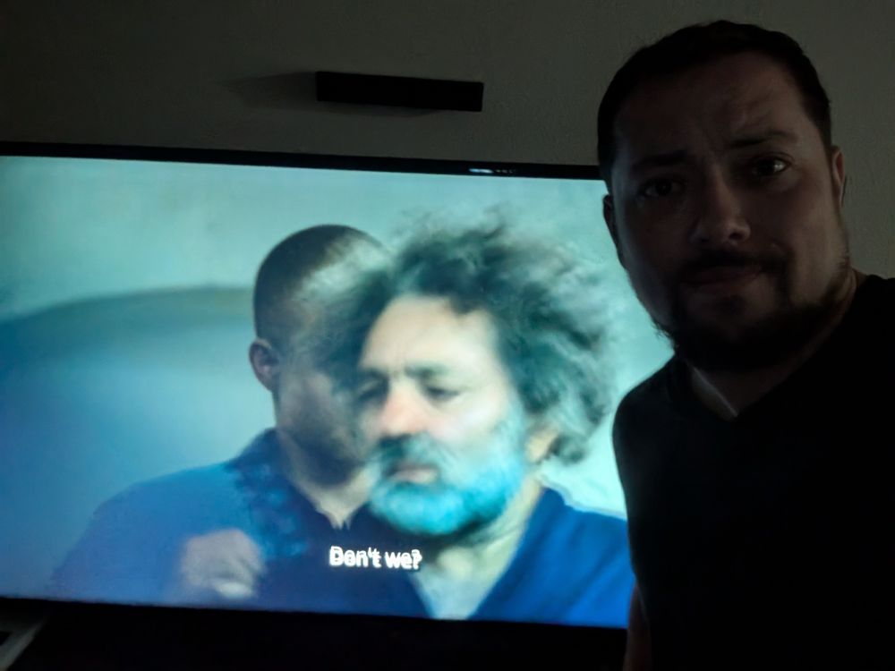 Me standing near my TV, which is playing a scene from The Wheel of Time, in which Rand (Josha Stradowski) puts a robe on Errol (Nasser Memarzia)