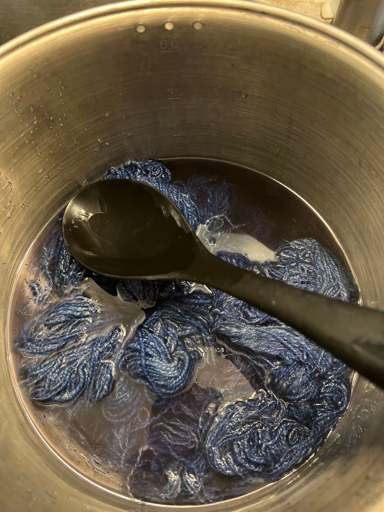 A stainless steel pot half filled with blue water and blue yarn. There is a large black plastic spoon in it
