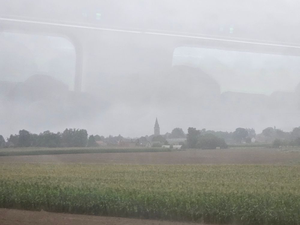 A flat landscape under a grey sky. On the horizon is a village. In the centre of the picture a church steeple reaches up to the sky.