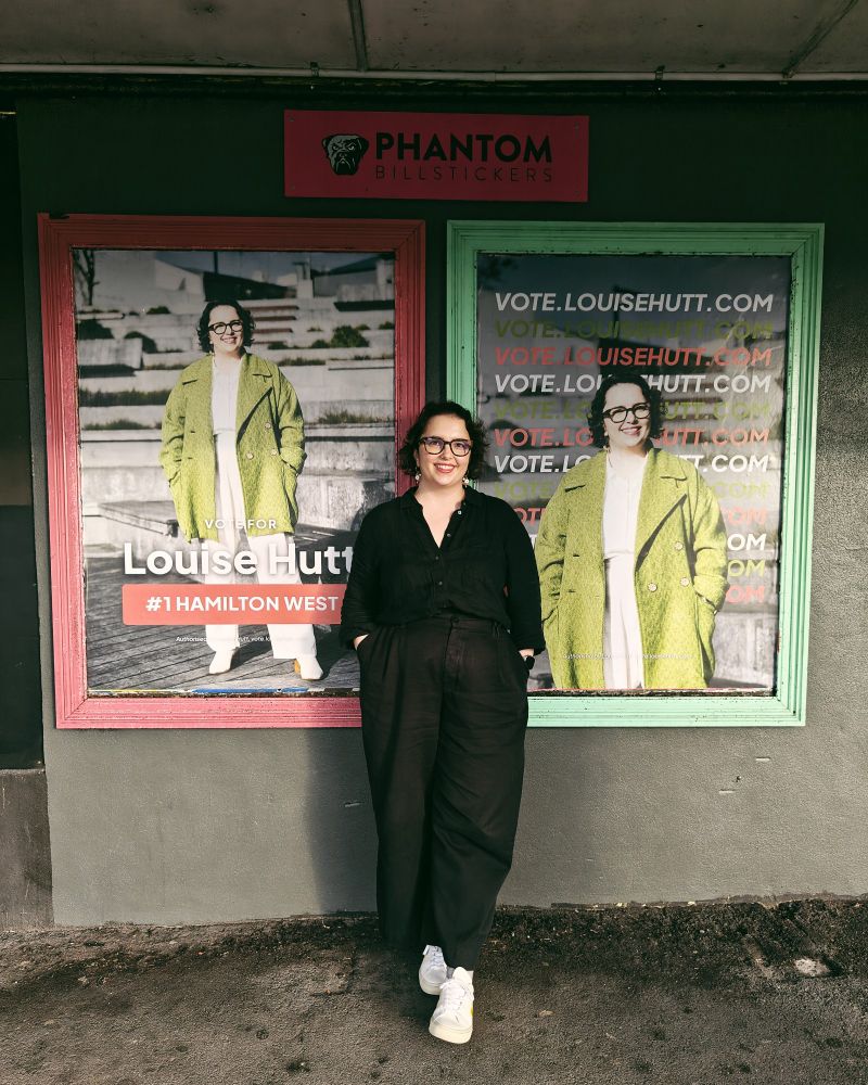 A photograph of Louise, standing in front of two posters - each saying a version of Vote Louise Hutt #1 Hamilton West