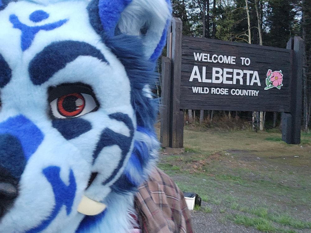 Blue sabertooth fursuit head in front of a sign that says "Welcome to Alberta."