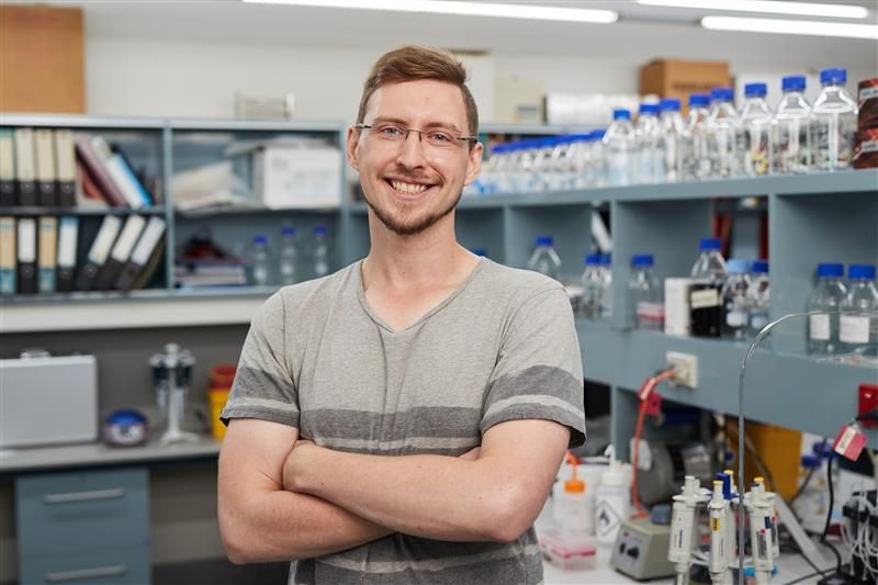 Person standing in a laboratory with arms crossed, surrounded by shelves containing bottles with blue caps, binders, and various scientific instruments.