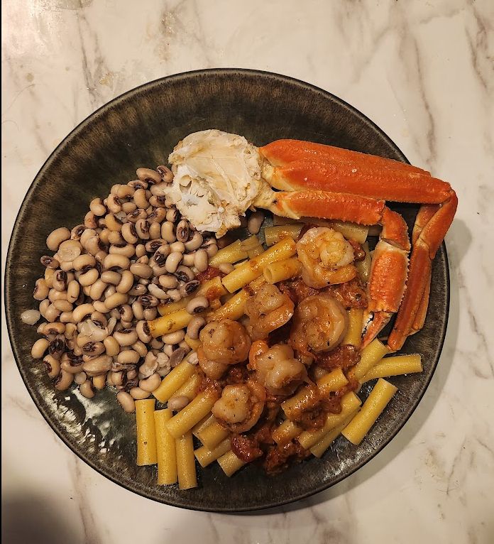 A plate of food consisting of Ziti pasta covered in a tomato pesto cream sauce topped with shrimp, a cluster of king crab legs, and black-eyed peas. 