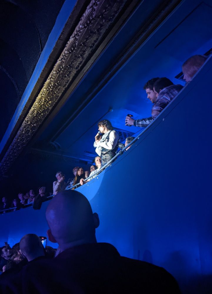 Spencer Sutherland, a man dressed in a sparkling waistcoat and trousers, holds a microphone, and stands amongst a crowd. A spotlight shines on him, while the people around watch on, bathed in blue light.