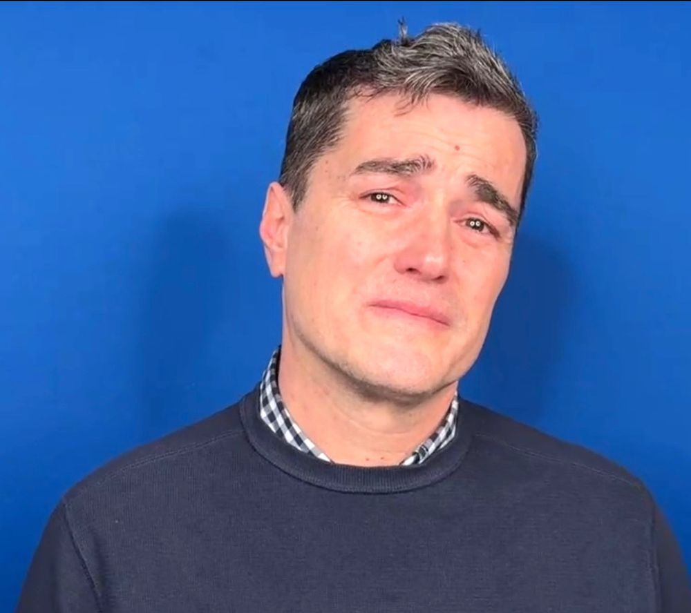 Brown haired man in front of a blue background looking very sad indeed