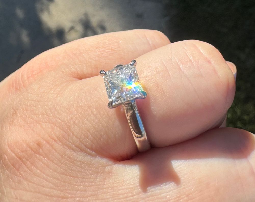 A close up of a left hand ring finger with a princess cut solitaire engagement ring on it, showing off a rainbow sparkle in the sunlight. 
