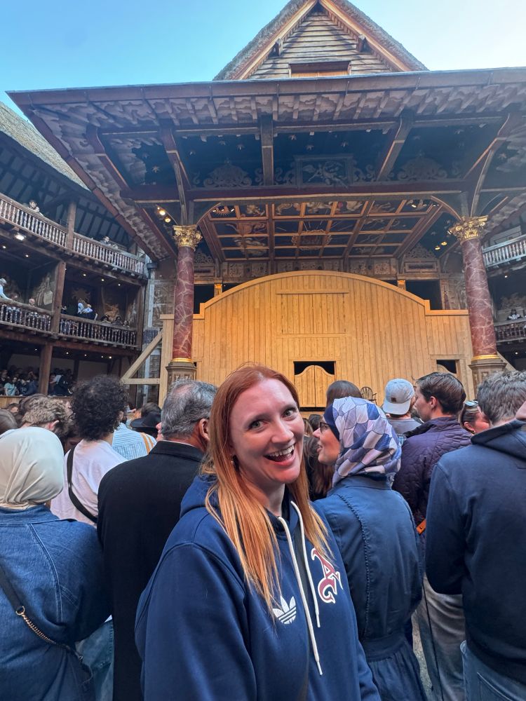 Allison, a ginger woman in a Navy Arsenal hoodie, is standing and smiling in the reconstructed Globe theatre in front of the iconic stage and surrounded by others in the crowd. 