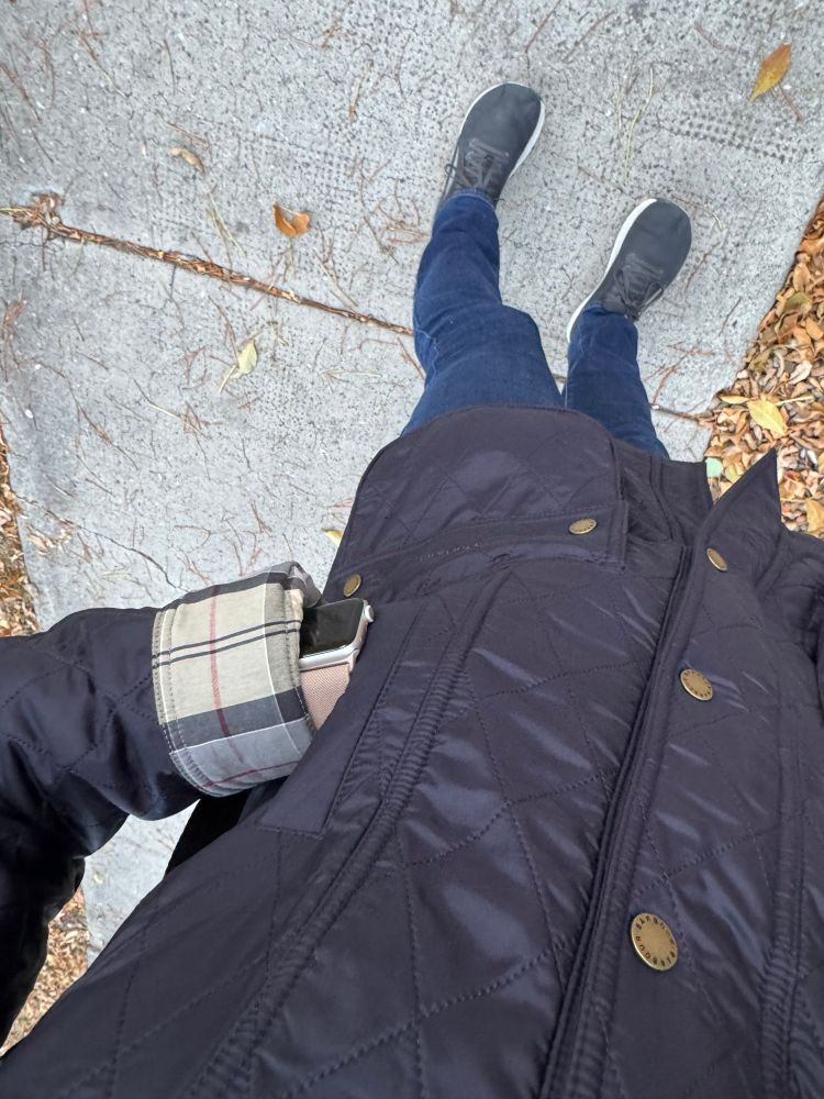 A woman’s torso, wearing a similar navy chore jacket. In the background, you can see a sidewalk and fallen leaves. The woman is wearing an Apple Watch and has her left hand in the warming pocket. 