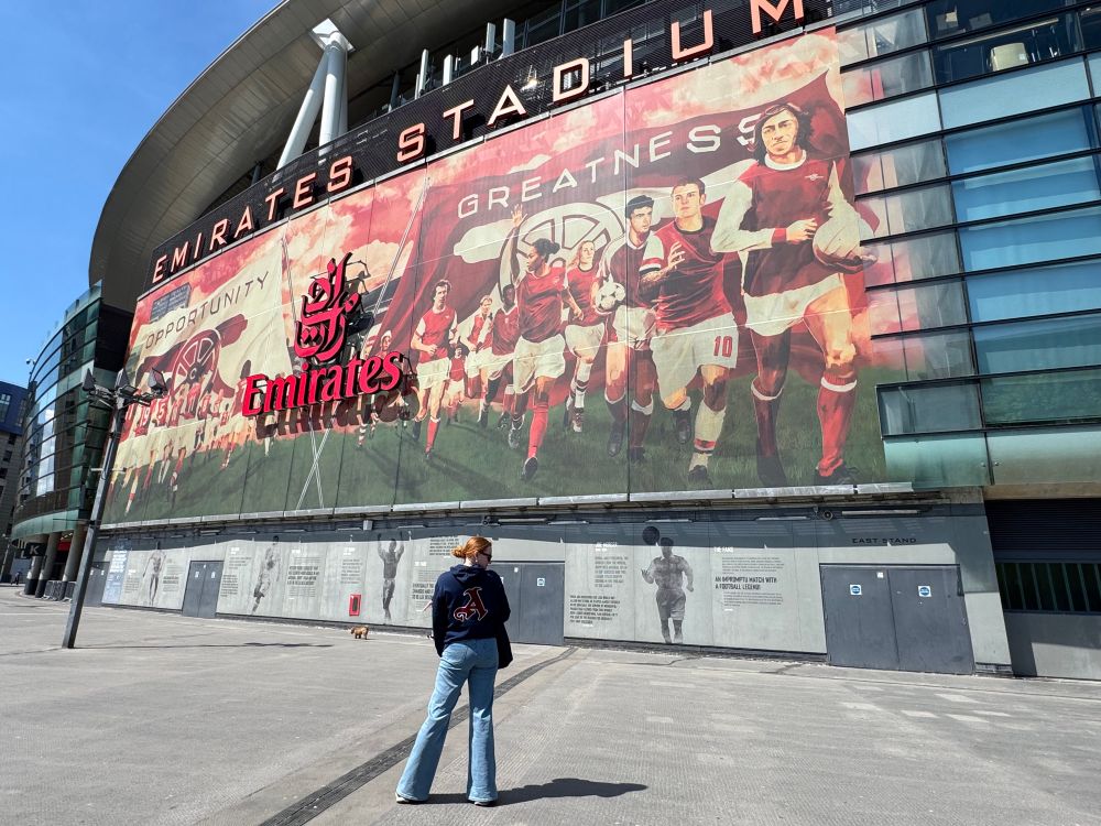 A ginger woman standing outside of Emirates Stadium wearing jeans and a navy hoodie with the Arsenal stylized “A” in maroon on the back 