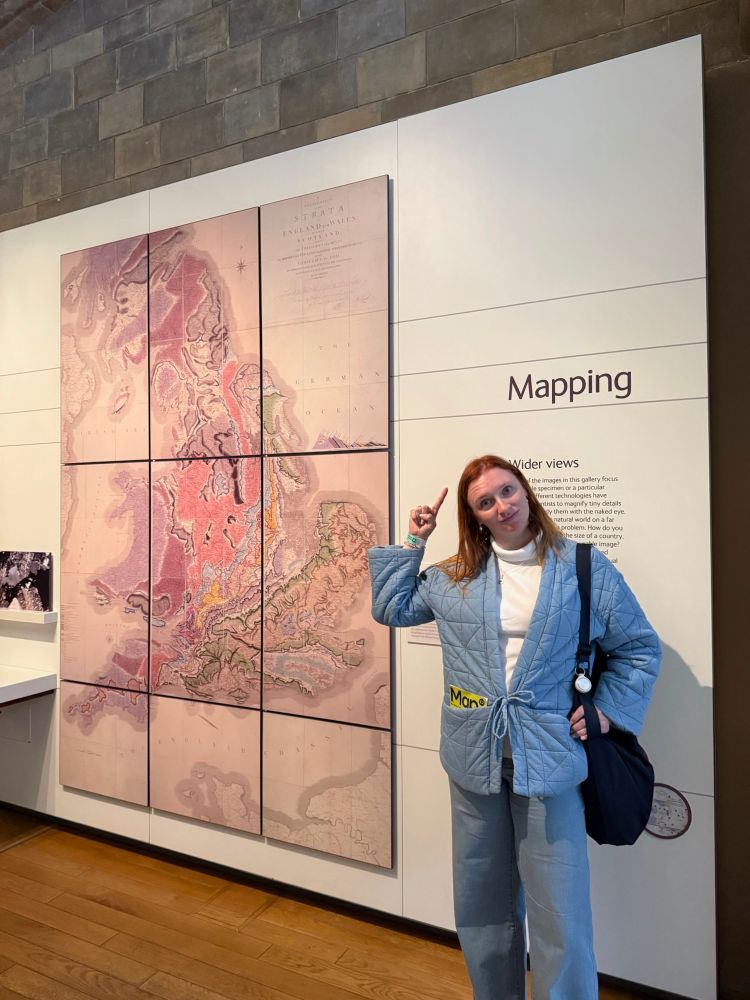 Allison at the British Natural History Museum standing beside a map, pointing to a sign that says “Mapping” above her, and with a map in her pocket. 