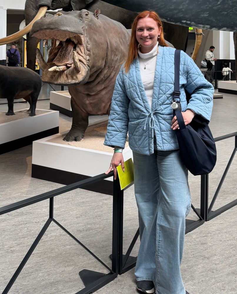 A woman in a blue quilted jacket stands in front of a life sized hippo model