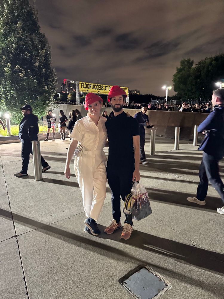 A woman in a full white jumpsuit stands with a man wearing all black. Both are wearing red Devo energy domes. 