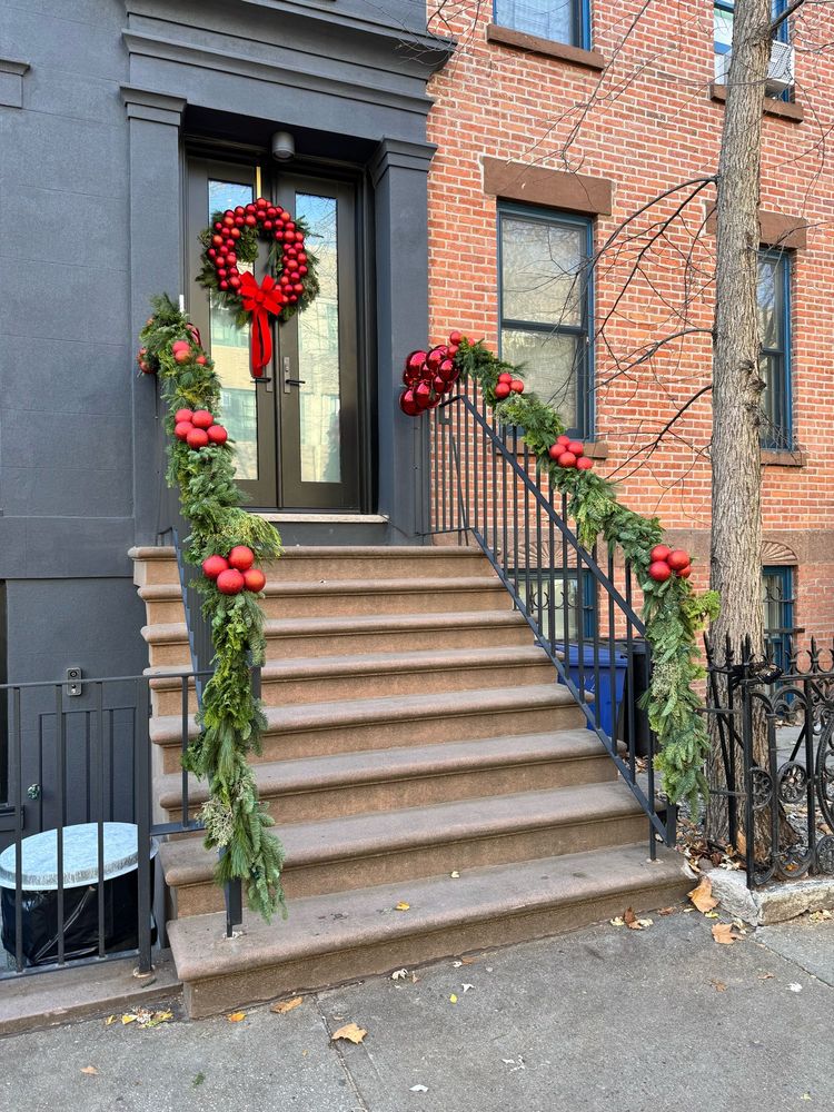 Entryway to townhouse in Brooklyn.  Front reps with decorated railings in pine tree greenery, large clusters of red ornament balls.  Grey building with large wreath on door also encircled with large red ornament balls and big red bow.  Attached house next door is exposed brick.  