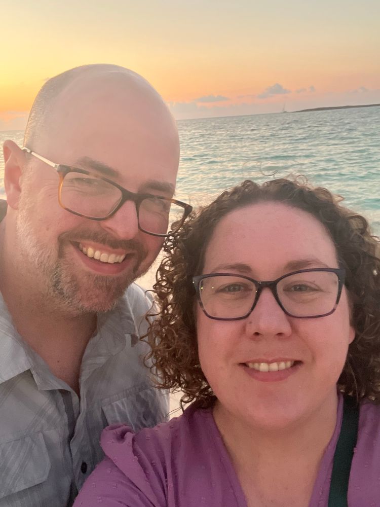 A smiling couple poses for a selfie at the beach during sunset. The man, on the left, has a shaved head, a trimmed beard, and is wearing glasses and a light gray shirt. The woman, on the right, has curly brown hair, is also wearing glasses, and has on a purple top. The ocean and a colorful sky with orange and pink hues are visible in the background.