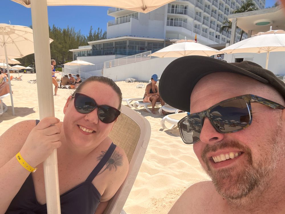 A smiling couple is relaxing on a sunny beach under a large umbrella. The woman is wearing dark sunglasses and a navy blue swimsuit, with a sunflower tattoo on her shoulder and a yellow wristband. The man is wearing a black cap and sunglasses, also smiling. Behind them are other beachgoers, white sand, lounge chairs, umbrellas, and a tall white resort building with balconies and palm trees in the background.