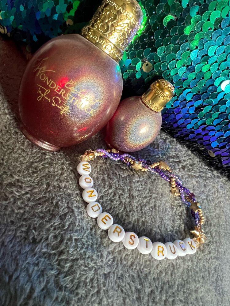 A purple and gold bracelet with Wonderstruck in gold letters next to a perfume bottle and a mini-bottle, both Wonderstruck by Taylor Swift. On a grey blanket with blue/green sequins behind. 