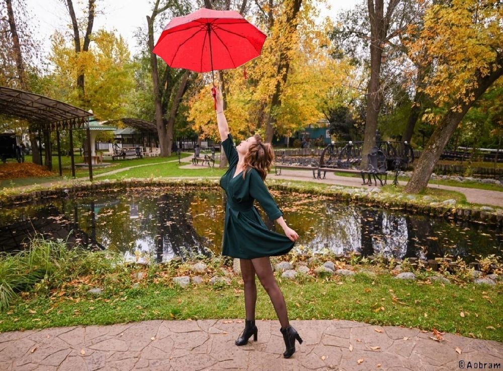 Photo of a woman seen from her left side standing before a pond in a park. She wears a mini dress, stockings and ankle boots. With a stretched right arm she upholds an unfolded red umbrella. On the background trees with autumn foliage reflected in the pond surface.