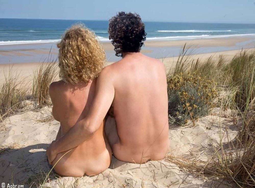 Foto van een naakte vrouw en naakte man op de rug gezien. Ze zitten dicht naast elkaar op een duintop. Ze kijken uit over het strand, de branding en de zee.