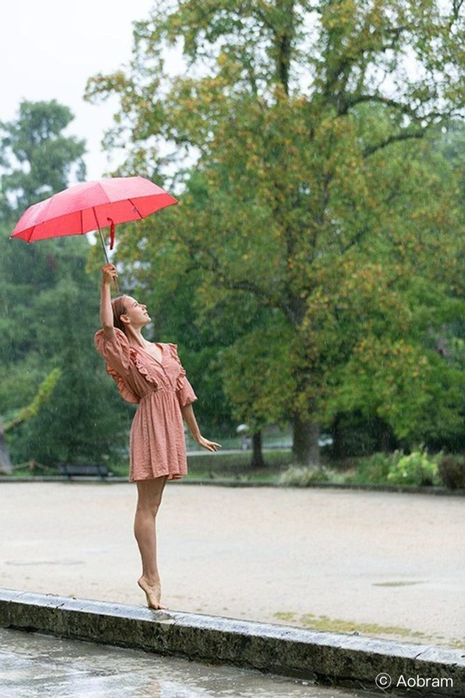 Photo of a woman, seen from beside, standing in a stretched pose barefoot on her toes, on a stone edge by a park. She wears a mini dress and upholds with one stretcher arm an unfolded red umbrella.