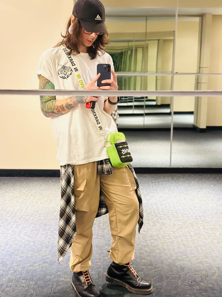 a full length mirror selfie of jay with a black adidas hat, white ghost and darkness shirt with a death moth, khaki joggers with a flannel tied around her waist and doc martens boots and an electric green varials hip back that says in darkness on the strap. her hair is parted in the middle and curling at her shoulders. shes holding a vape in her hand, and most of her sleeve can be seen in the mirror